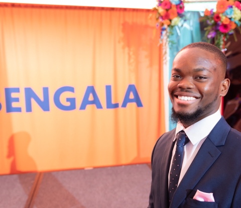 Smiling student in suit and tie standing in front of orange curtain backdrop that reads BENGALA