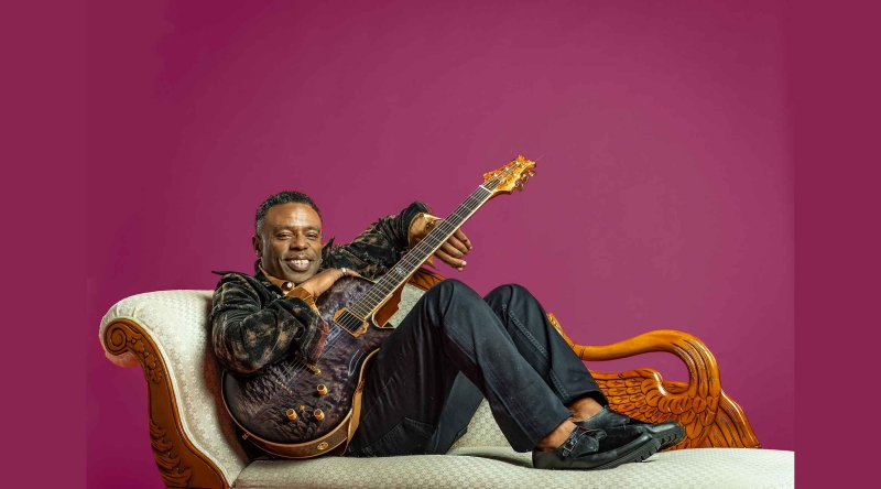 Norman Brown holding a guitar and reclining on a chaise longue